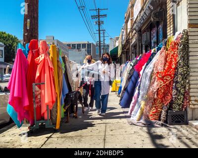 Bulloni di tessuti sono in vendita sul marciapiede di Il quartiere degli indumenti di Los Angeles come una coppia che indossa il viso le maschere a causa della pandemia del coronavirus trasportano un Foto Stock