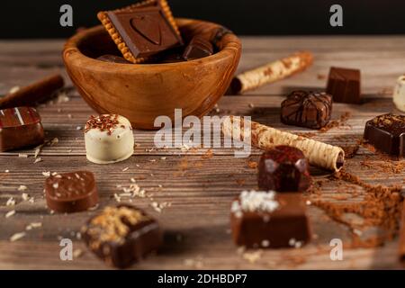 Una ciotola di cioccolatini assortiti e biscotti al cioccolato su sfondo di legno con bastoncini di cacao in polvere alla cannella, pirouettes e cocco spruzzano la creazione Foto Stock