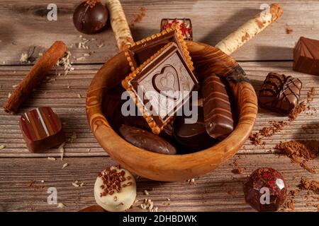 Una ciotola di cioccolatini assortiti e biscotti al cioccolato su sfondo di legno con bastoncini di cacao in polvere alla cannella, pirouettes e cocco spruzzano la creazione Foto Stock