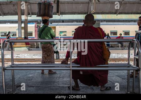 Yangon, Myanmar - 31 dicembre 2019: Un monaco in tunica rossa seduto su una semplice panchina d'acciaio presso la stazione ferroviaria circolare di Yangon Foto Stock