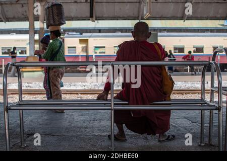 Yangon, Myanmar - 31 dicembre 2019: Un monaco in tunica rossa seduto su una semplice panchina d'acciaio presso la stazione ferroviaria circolare di Yangon Foto Stock