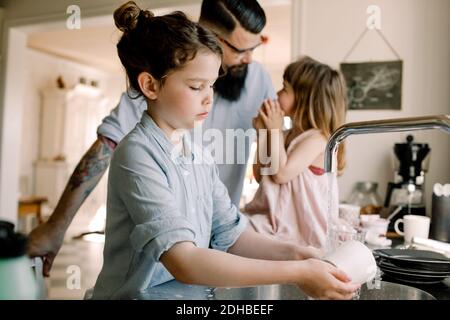 Ragazza che lavava la tazza al lavandino mentre il padre parla con la figlia in cucina Foto Stock