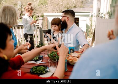 Famiglia multi-generazionale che ha cibo mentre le figlie usano la tecnologia nel patio Foto Stock