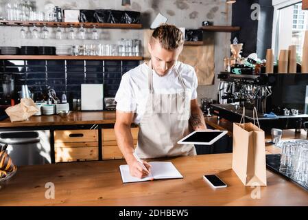 giovane cameriere che scrive nel notebook mentre tiene il tablet digitale vicino smartphone e borsa di carta Foto Stock