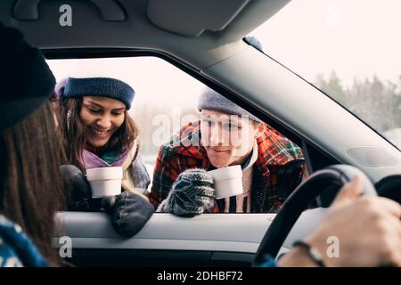 Uomo sorridente e donna che parlano con un amico seduto in macchina mentre si ha il caffè durante l'inverno Foto Stock