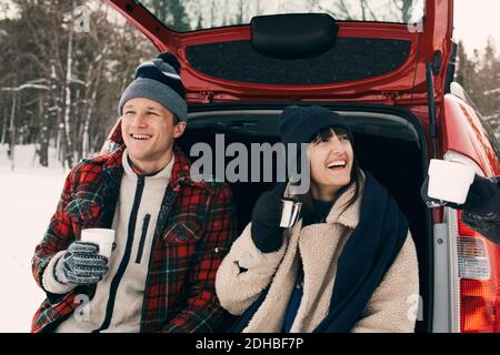 Donna felice che ha caffè mentre si siede con gli amici in auto bagagliaio durante l'inverno Foto Stock