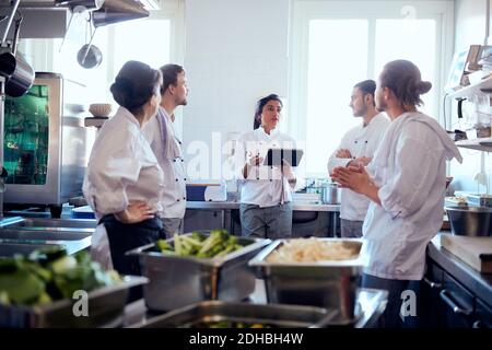 Cuoco femmina medio adulto che tiene un tablet digitale mentre discute con squadra in cucina Foto Stock