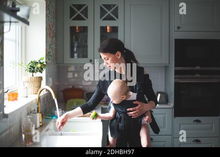 Madre lavando la mano nel lavandino della cucina mentre trasportano la bambina a casa Foto Stock