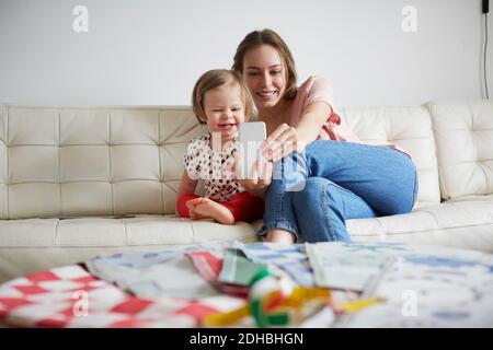 Sorridente madre e figlia che prendono selfie attraverso smartphone mentre seduto sul divano a casa Foto Stock