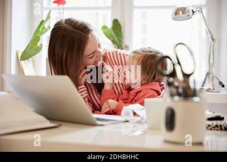 Donna sorridente che parla con la figlia mentre lavora su un computer portatile a. casa Foto Stock