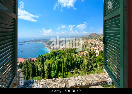 Vista attraverso una finestra aperta con persiane della costa e del villaggio di Taormina Italia, sull'isola della Sicilia nel Mar Mediterraneo Foto Stock