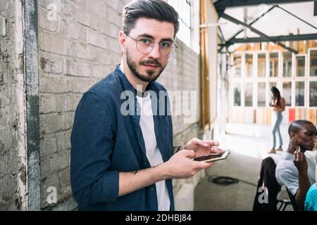 Ritratto di giovane uomo d'affari sicuro in piedi con smartphone contro muro di mattoni in un luogo di lavoro creativo Foto Stock