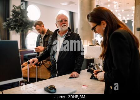Uomo anziano che guarda lontano mentre si alza in piedi con il receptionist alla reception in hotel Foto Stock