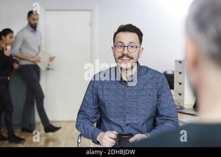 Sorridente uomo d'affari che tiene la tazza del caffè mentre discute con il professionista a. ufficio Foto Stock