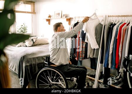 Vista laterale di un uomo maturo per disabili che appende una t-shirt bianca portabiti a casa Foto Stock