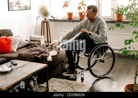 L'intera lunghezza di uomo maturo disabile che indossa scarpe mentre si siede su sedia a rotelle nel soggiorno di casa Foto Stock