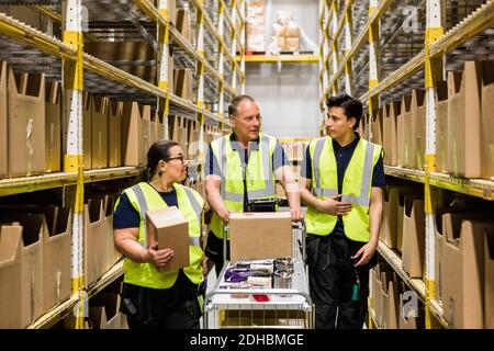 I colleghi multietnici discutono mentre camminano con il carrello sulla navata in mezzo rack nel magazzino di distribuzione Foto Stock