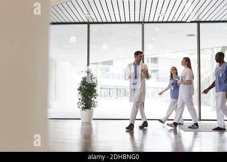 Tutta la lunghezza dei medici e degli infermieri che discutono mentre camminano dentro corridoio contro la finestra dell'ospedale Foto Stock