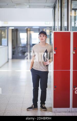 Ritratto di ragazzo fidato tenente libro mentre si sta in piedi armadietti nel corridoio della scuola superiore Foto Stock