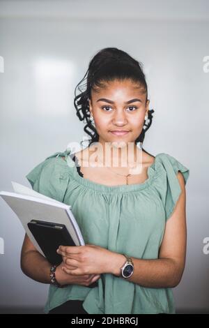 Ritratto di fidata ragazza adolescente in piedi con libro contro lavagna bianca in laboratorio informatico presso la scuola superiore Foto Stock