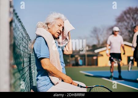 Donna anziana strofinata faccia con asciugamano mentre si siede a. campo da tennis Foto Stock