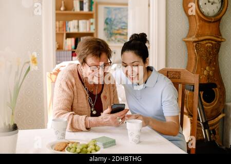 Sorridente giovane volontaria femminile che assiste anziana donna in pensione utilizzando Smart telefono al tavolo in casa di cura Foto Stock