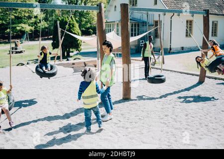 Insegnanti in piedi mentre gli studenti si divertono a swing nel parco giochi Foto Stock