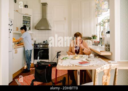 Donna anziana in pensione che mangia la colazione mentre la donna si prende cura della cucina a casa di cura Foto Stock