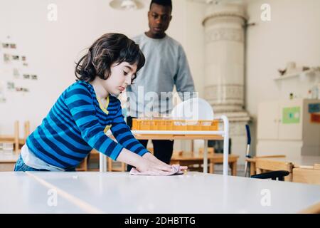 Tavolo per la pulizia del ragazzo con il panno per piatti mentre l'insegnante sta in piedi scuola presso l'asilo Foto Stock