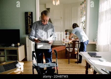 Uomo anziano pensionato che legge giornale mentre caregiver femminile che lavora dentro cucina a casa di cura Foto Stock