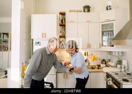 Uomo anziano in pensione che mostra lo smartphone al lavaggio volontario femminile piatti in cucina a casa di cura Foto Stock