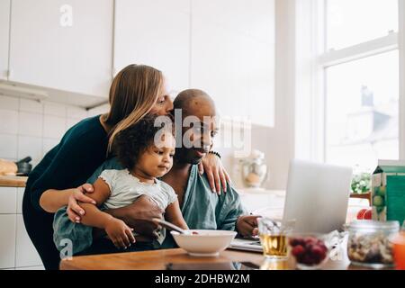 Donna che abbracciava e baciava l'uomo con la figlia usando il laptop a. casa Foto Stock