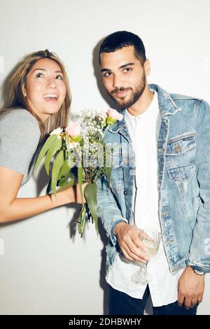 Donna allegra che dà fiori al giovane uomo in piedi con una bevanda contro la parete bianca a casa Foto Stock