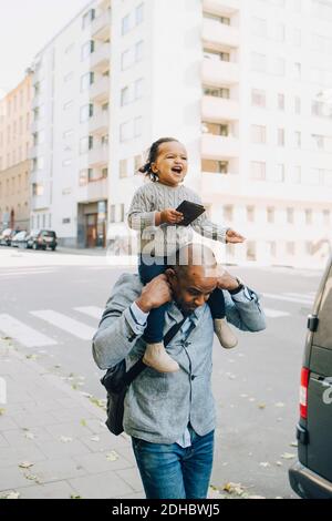 Padre che porta con sé una figlia allegra che tiene lo smartphone mentre si cammina sentiero in città Foto Stock