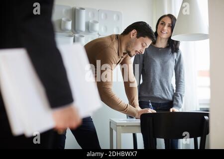 Donna che guarda l'agente immobiliare mentre l'uomo firma i documenti a casa nuova Foto Stock