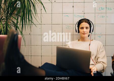 Ritratto di giovane e sicuro hacker di computer che ascolta con le cuffie mentre si è seduti con il computer portatile contro la piastrella w Foto Stock