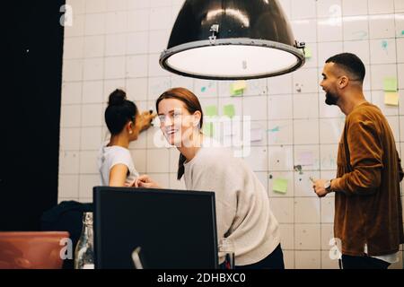 Sorridente giovane donna d'affari che guarda via mentre gli hacker progettano sopra la parete in un piccolo ufficio Foto Stock