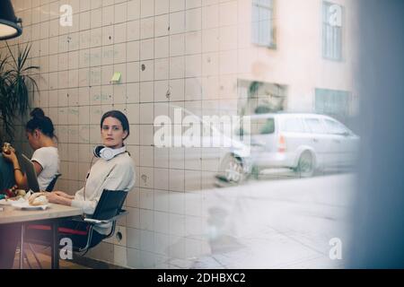 Giovane donna d'affari che guarda via mentre si siede alla scrivania in piccolo ufficio visto attraverso la finestra Foto Stock