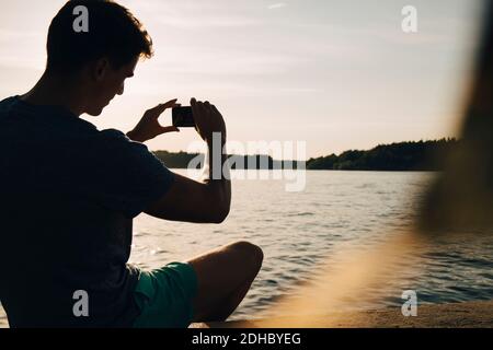 Giovane uomo che fotografa il lago mentre si siede al molo durante l'estate Foto Stock