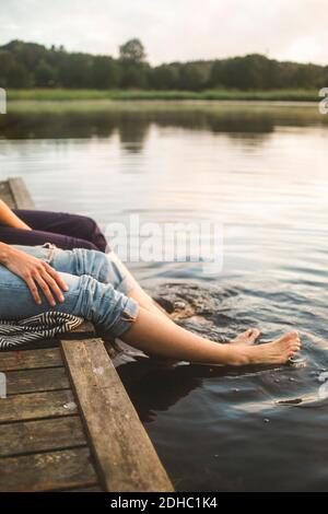 Sezione bassa di amici femminili che penzolano le gambe nel lago mentre seduto sul molo Foto Stock