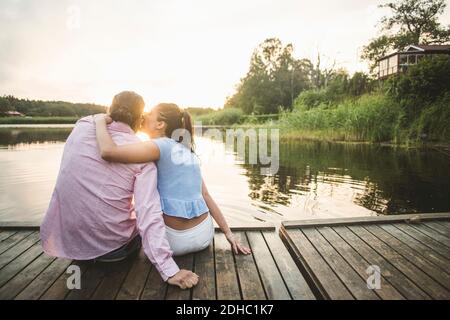 Vista posteriore della fidanzata baciante seduta con le braccia intorno sul molo sopra il lago durante il tramonto Foto Stock