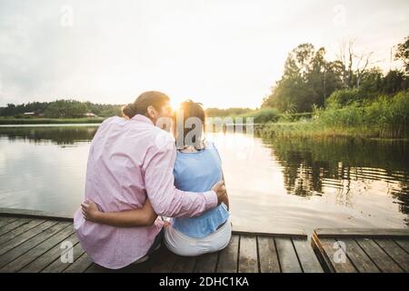 Vista posteriore della fidanzata baciante seduta con le braccia intorno sul molo sopra il lago durante il tramonto Foto Stock