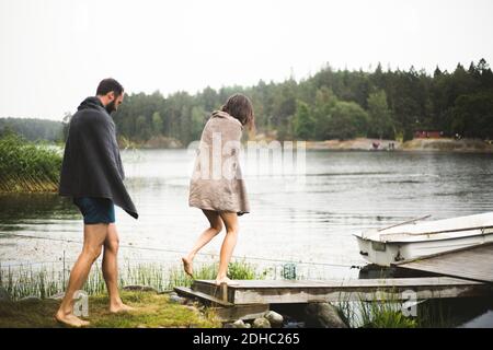 Amici maschili e femminili avvolti in asciugamani camminando verso il molo sul lago durante la fuga del fine settimana Foto Stock