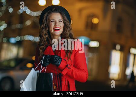 Winter Fun. Donna di 40 anni felice elegante shopper in cappotto rosso e berta nera con borse per lo shopping fuori in città strada in serata. Foto Stock