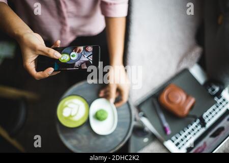 Sezione centrale del blogger femminile fotografando il tè matcha e snack attraverso smartphone in ufficio creativo Foto Stock