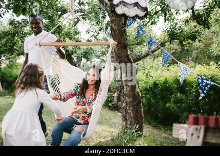 Padre che spinge l'amaca mentre donna felice che tiene la mano della figlia in cortile Foto Stock