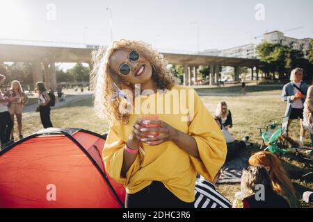 Ritratto di donna sorridente con drink in piedi contro gli amici a. evento musicale estivo Foto Stock