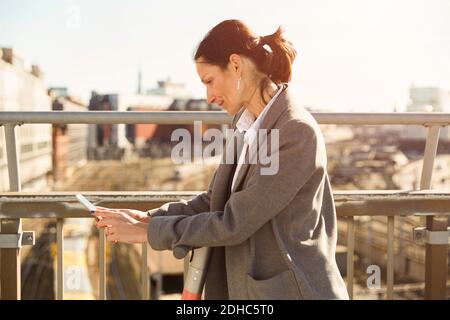 Vista laterale di una donna matura che utilizza uno smartphone mentre si è in piedi con scooter elettrico a spinta sul ponte in città Foto Stock