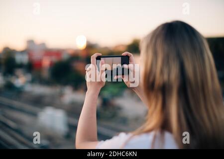 Giovane donna che fotografa Fernsehturm attraverso lo smartphone durante il tramonto Foto Stock