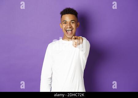 Immagine di bel giovane afroamericano in felpa bianca, puntando il dito verso la macchina fotografica e sorridendo, scegliendo voi, in piedi Foto Stock